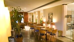 Comedor de hotel con mesas y sillas de madera, plantas en macetas y un muro divisor de vidrio.