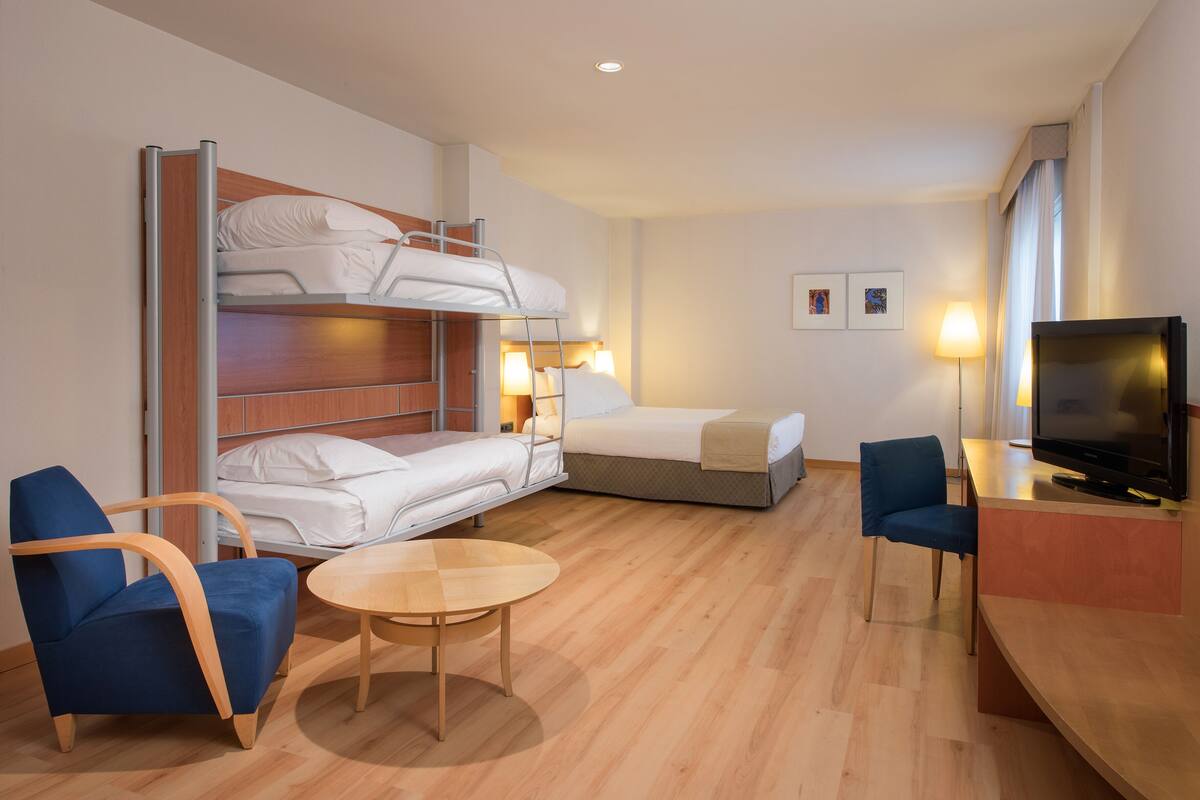 A hotel room with bunk beds, a desk with a TV, two blue chairs, a round table, and two bedside lamps.