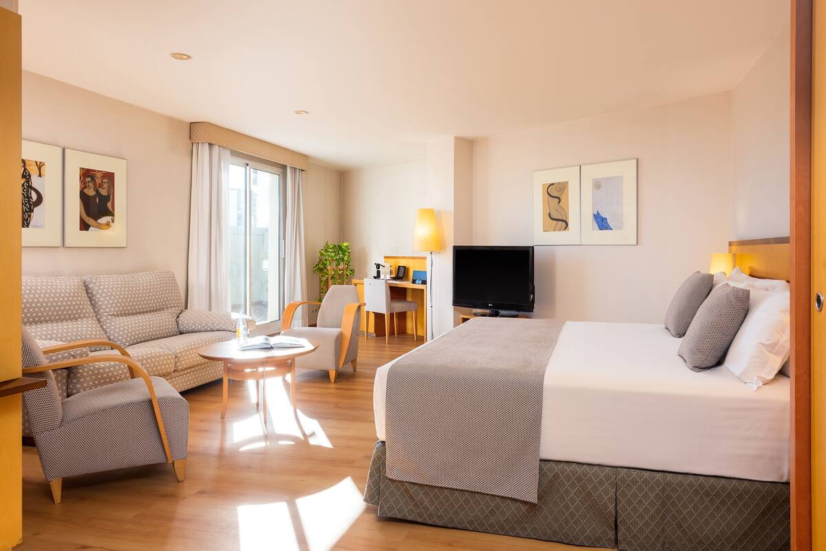 A hotel room with a bed, a sofa, a chair, a desk with a lamp, and a TV.