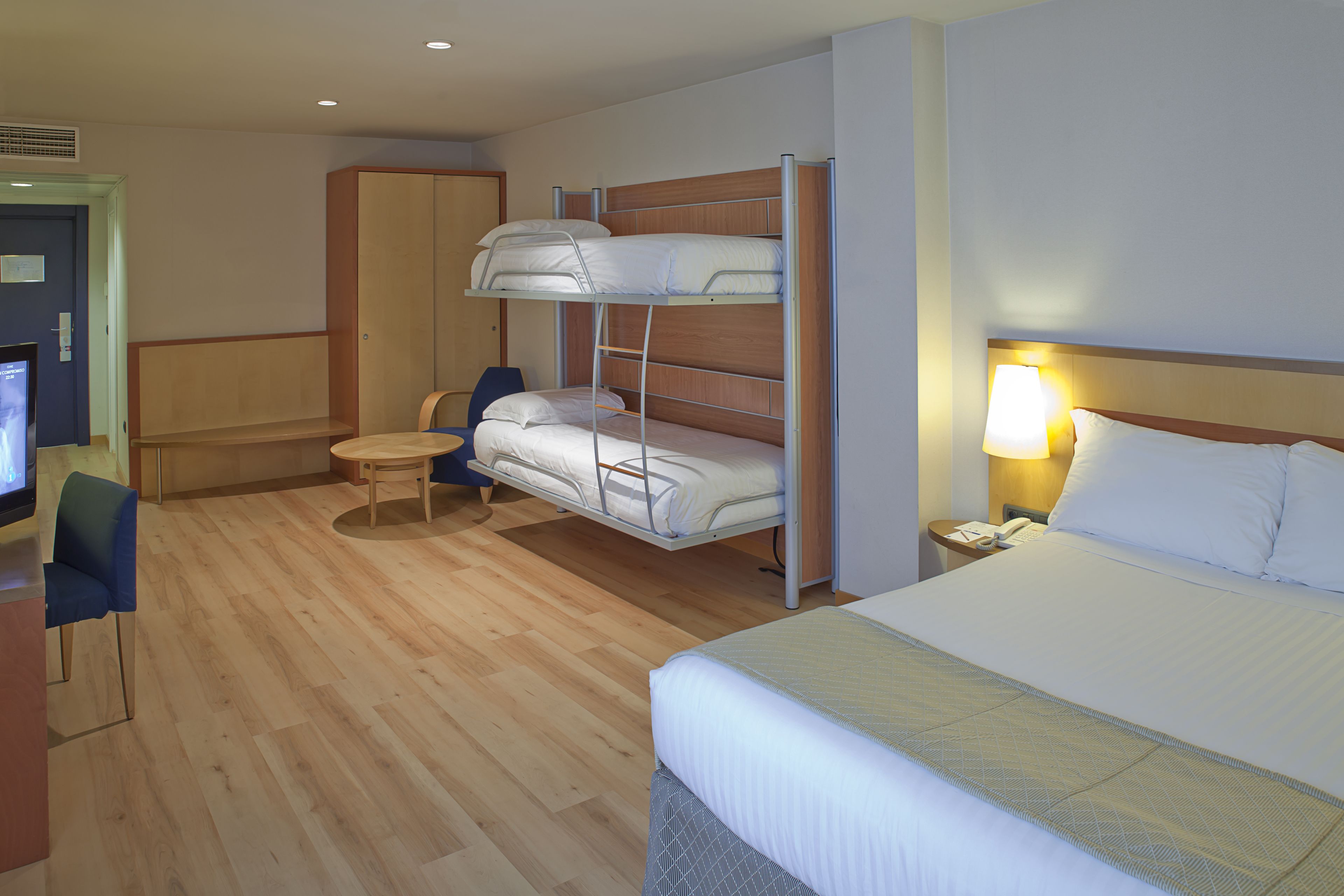 A hotel room with a bunk bed, a single bed, a desk with a chair, a TV, and a bedside lamp.