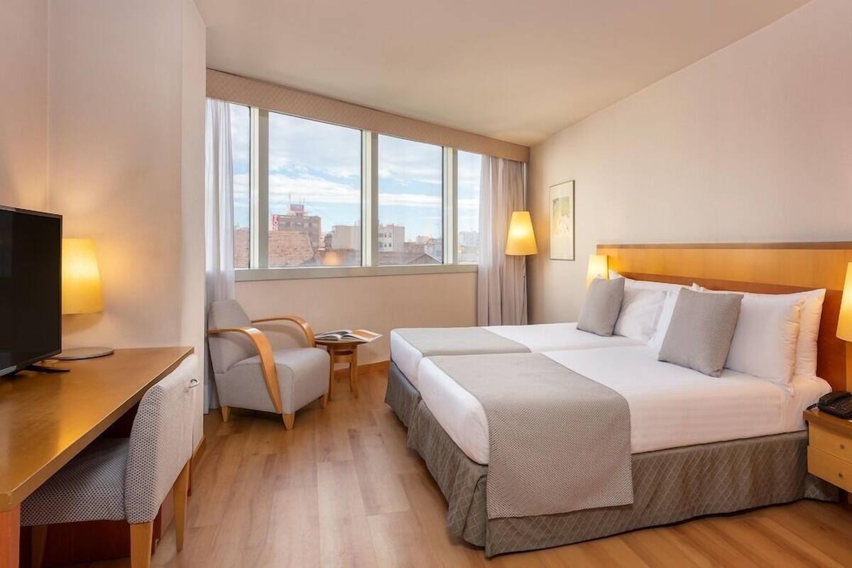 A hotel room with a bed, a desk, a TV, and a window with a city view.