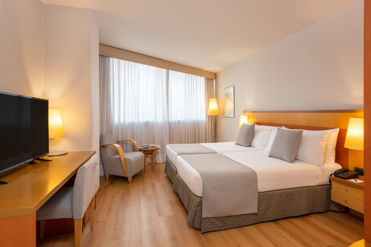 A hotel room with a bed, bedside table, lamp, TV, and a desk with a chair.