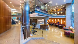 A modern hotel lobby with a central reception desk, multiple seating areas, and a staircase leading to upper floors. - Hotel Castellon Center Affiliated by Meliá (Castelló de la Plana)