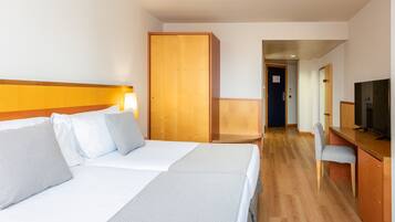 A hotel room with two beds, a wooden wardrobe, a desk with a chair, and a television.