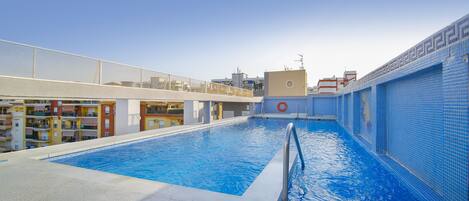 Una piscina con agua cristalina azul, rodeada por un deck de baldosas blancas y una pared de azulejos azules.