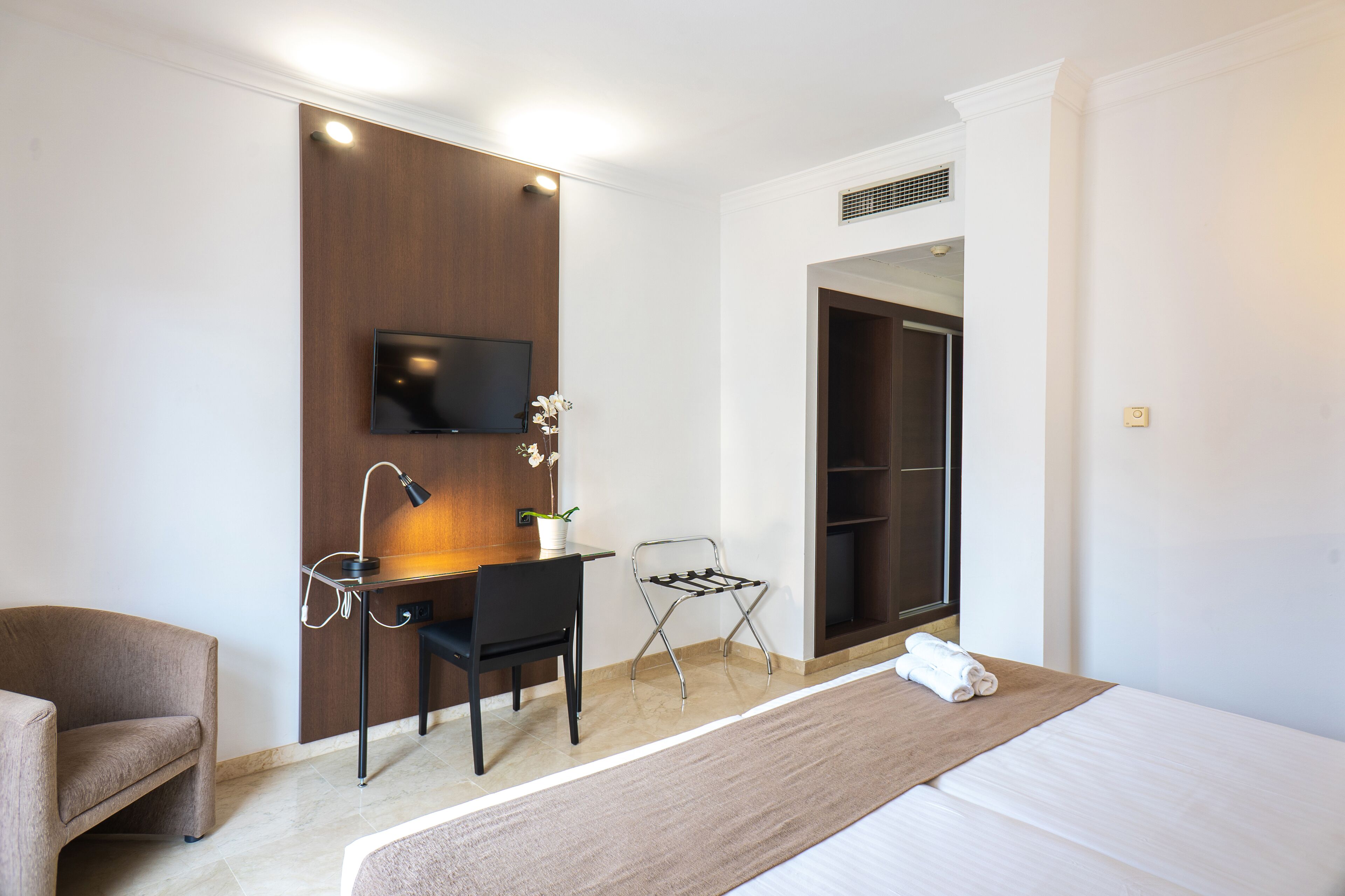 Habitación de hotel con cama, escritorio y silla, televisor montado en la pared, una planta pequeña en maceta y un espejo.