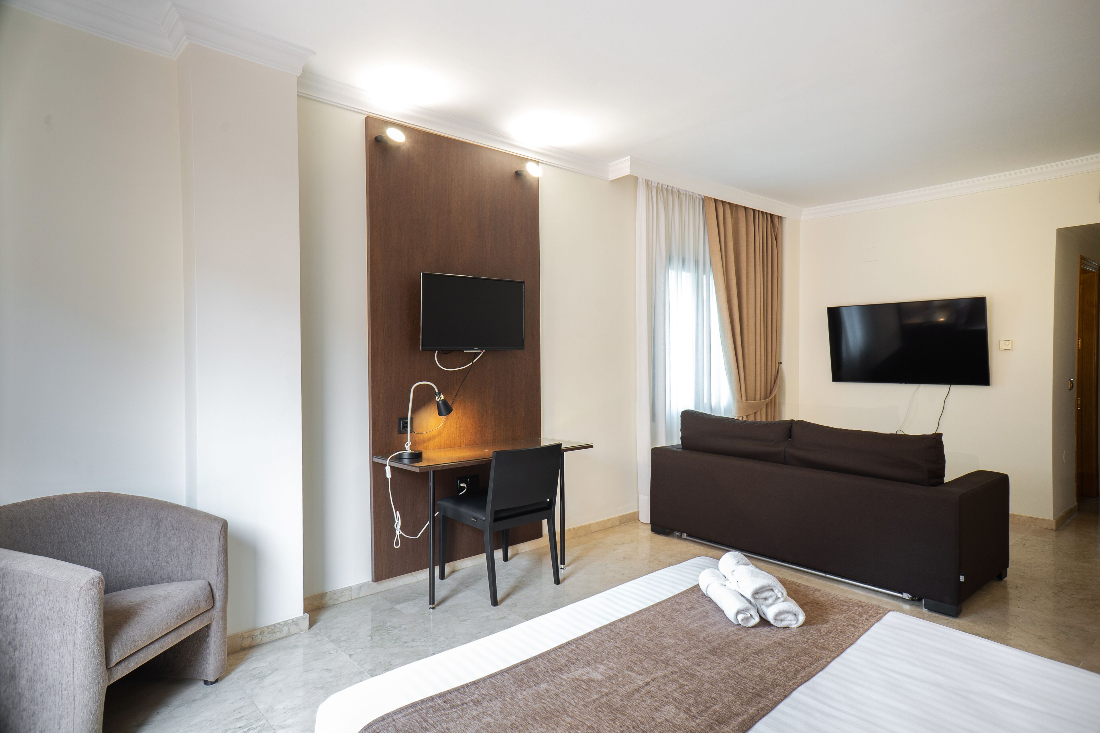 A hotel room with a bed, a desk with a chair, a sofa, and two TVs mounted on the walls.