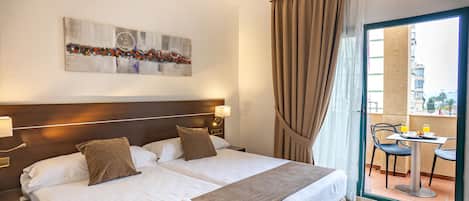 Habitación de hotel con cama doble, cabecero, mesita de noche con lámpara, ventana con cortinas y vista al balcón exterior.