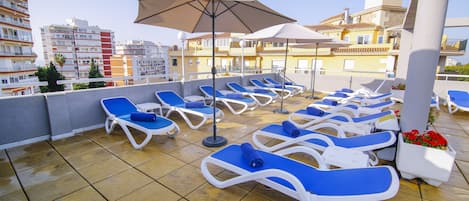 A rooftop terrace with blue lounge chairs and umbrellas, overlooking a cityscape with buildings and a tower.