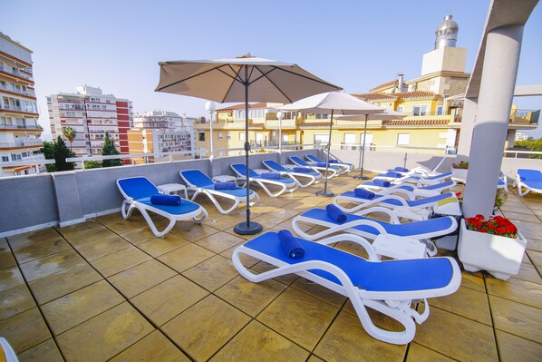 A rooftop terrace with blue lounge chairs and umbrellas, overlooking a cityscape with buildings and a tower.