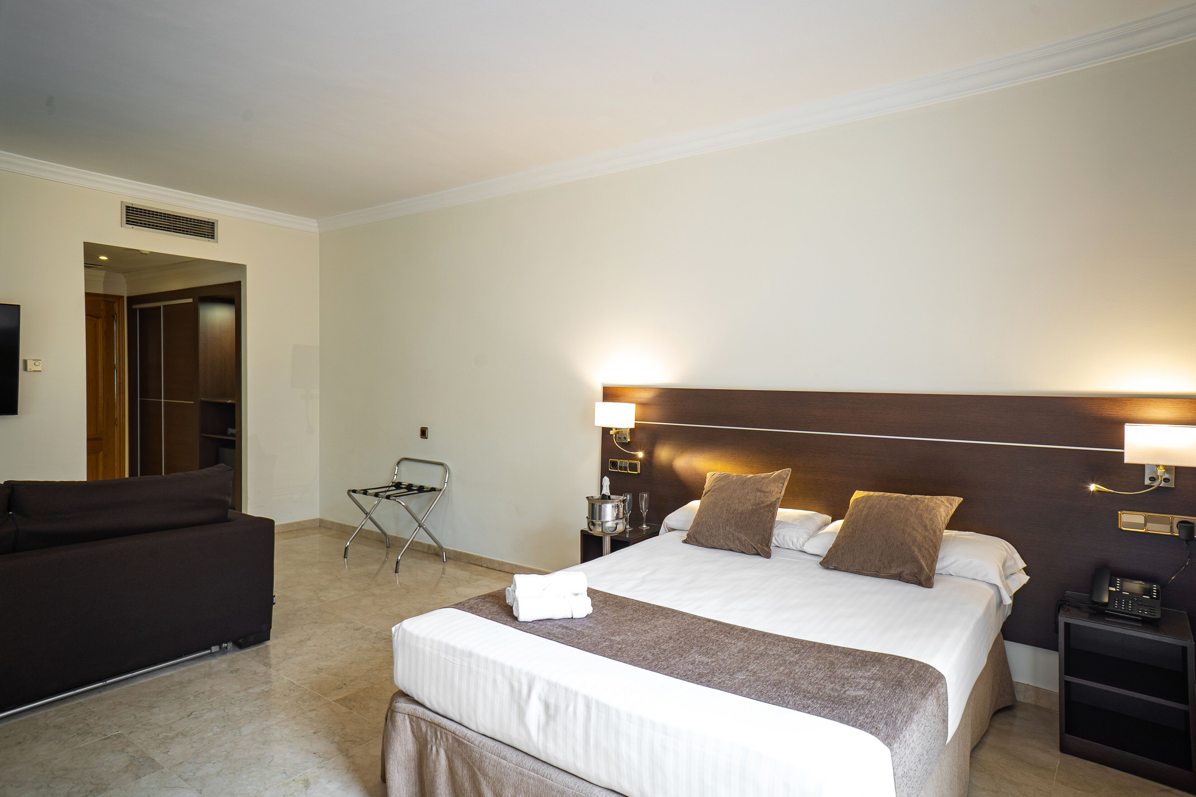 A hotel room with a large bed, a dark-colored sofa, a small table with a lamp, and a chair.