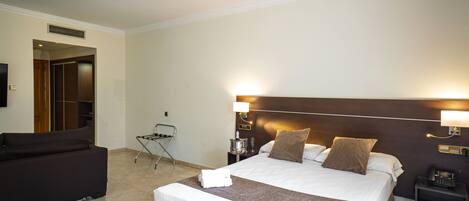 A hotel room with a large bed, a dark-colored sofa, a small table with a lamp, and a chair.