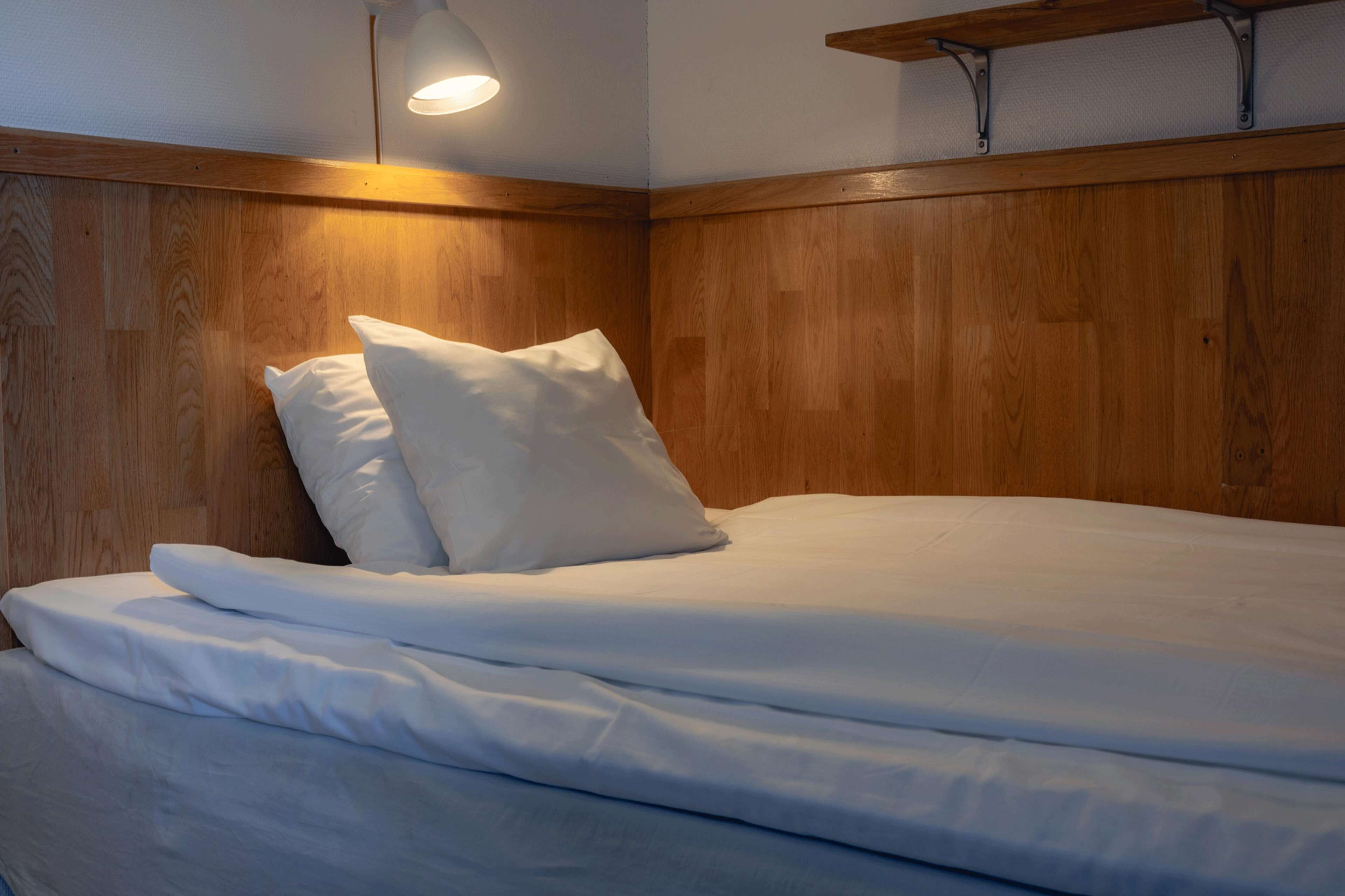 Una cama bien hecha con sábanas y almohadas blancas, cabecera de madera y una estantería en la pared.