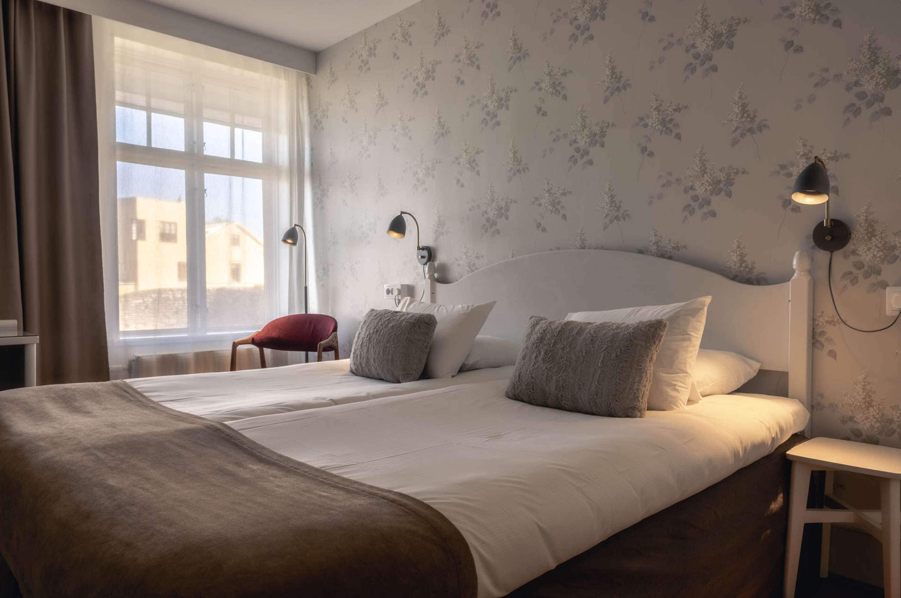 Une chambre d’hôtel avec un grand lit, deux lampes de chevet, un fauteuil rouge et une fenêtre donnant sur des immeubles.