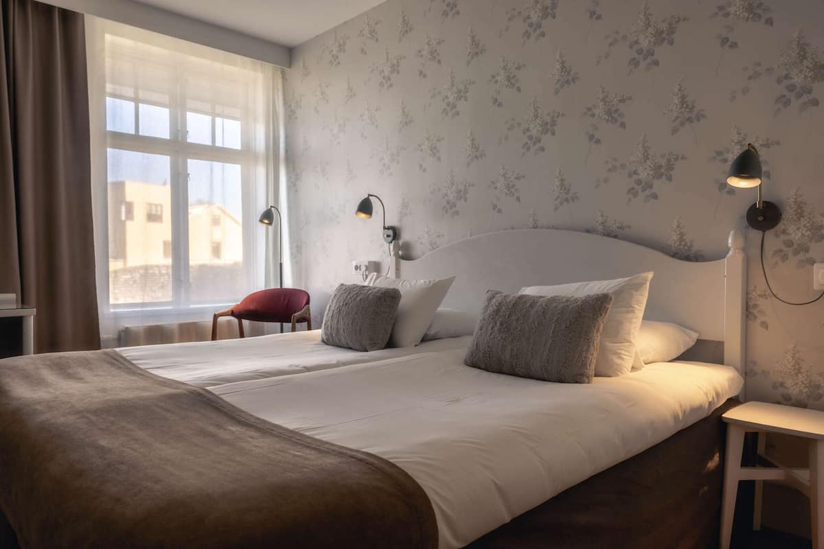 Une chambre d’hôtel avec un grand lit, deux lampes de chevet, un fauteuil rouge et une fenêtre donnant sur des immeubles.