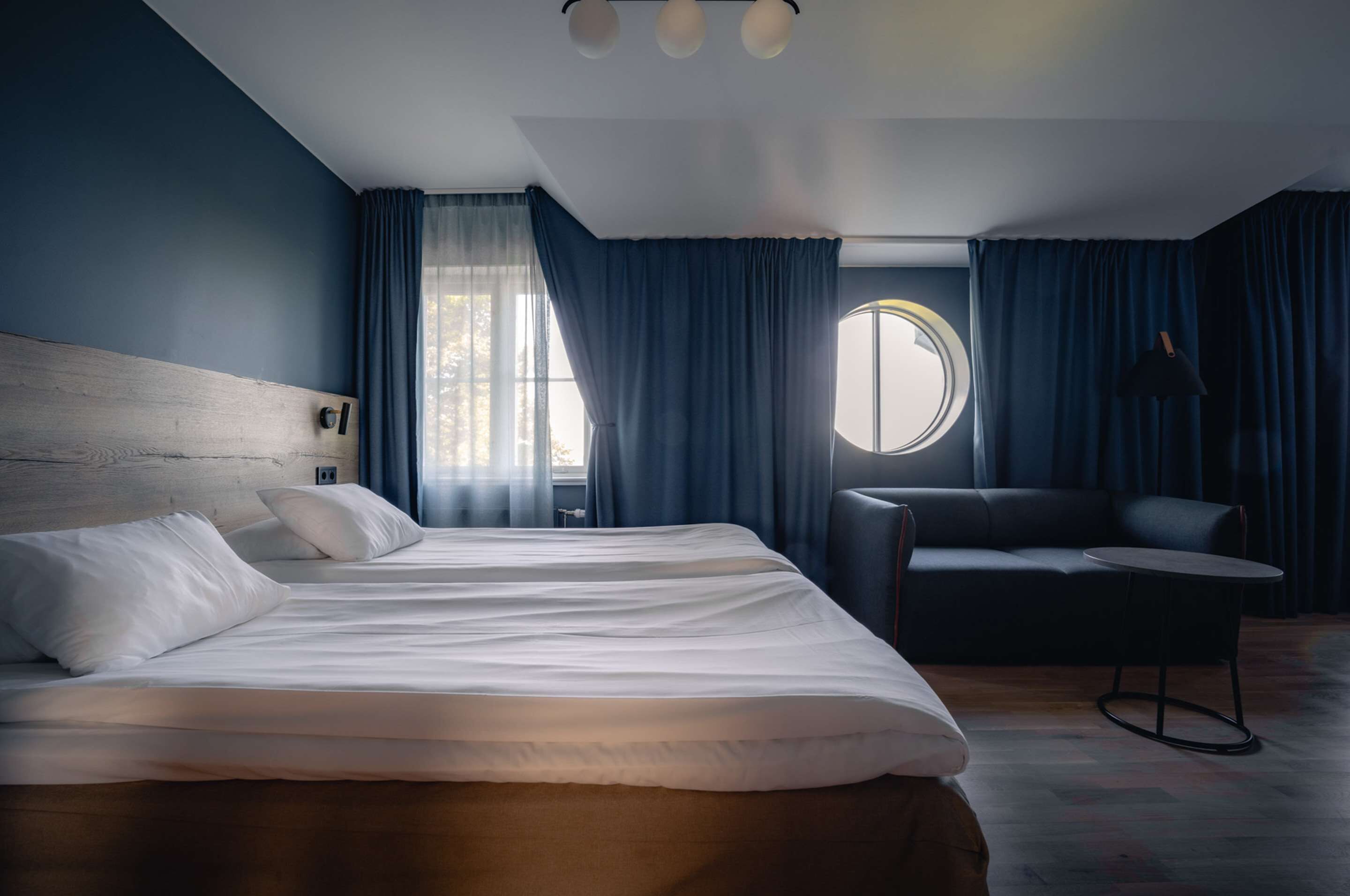 A modern hotel room with a large bed, a round window, a sofa, and a small table.