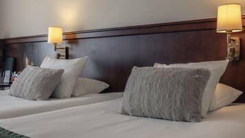 Habitación de hotel con dos camas, cada una con ropa blanca y mullidos almohadones grises. A cada lado de las camas hay lámparas fijadas a la pared.