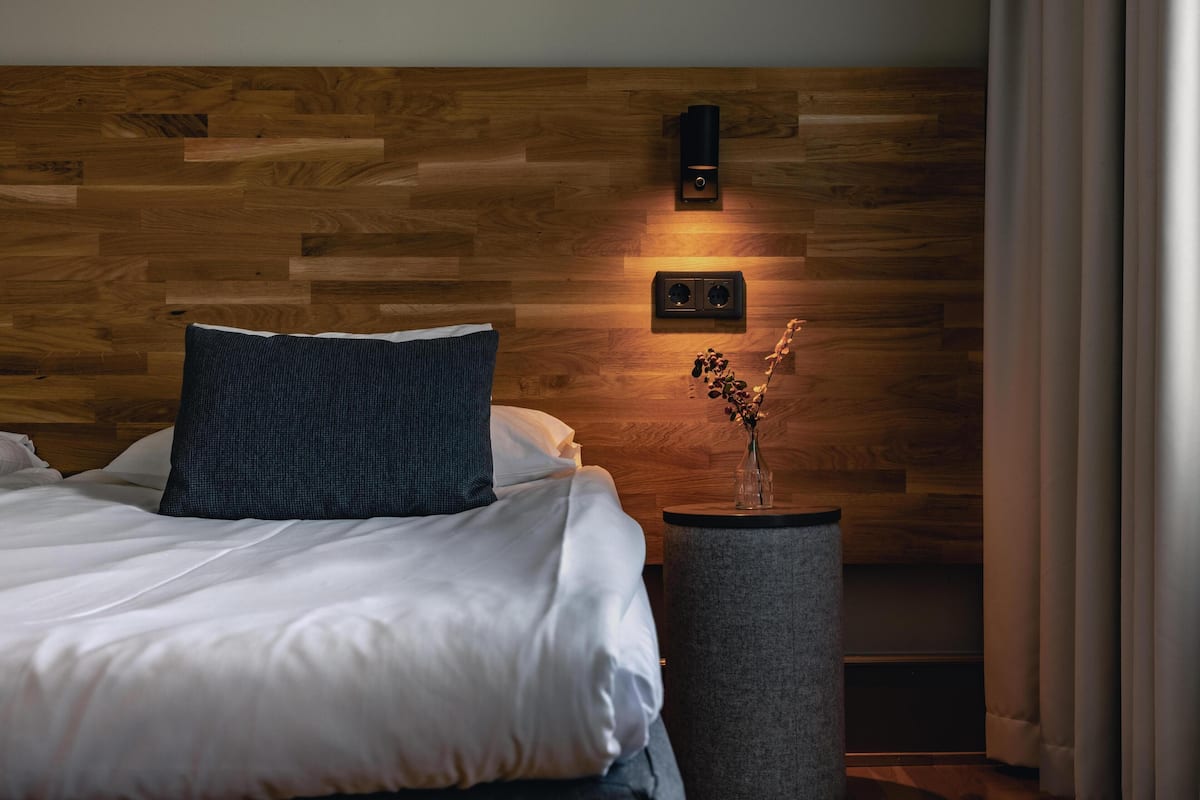 A bedroom with a wooden headboard, a bedside table with a vase, and a wall-mounted light fixture.