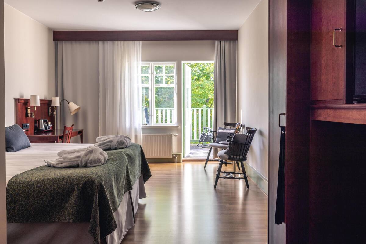 A hotel room with a bed, two bedside tables, a desk with a chair, a window with curtains, and a view of greenery outside.