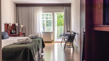 A hotel room with a bed, two bedside tables, a desk with a chair, a window with curtains, and a view of greenery outside.