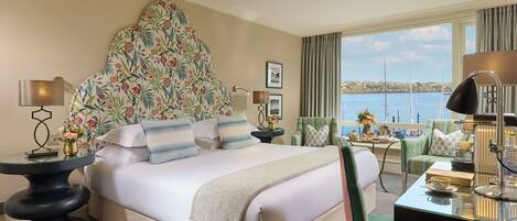 A hotel room with a large bed, a view of the sea, a desk with a lamp, and a chair.