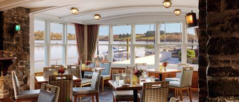 Un restaurant offrant une vue sur un plan d’eau et des bateaux, avec des tables et des chaises en bois, et des murs en pierre.