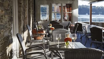 Un espace de restauration en plein air avec des chaises et des tables en osier, une vue sur l’eau et un mur en pierre.