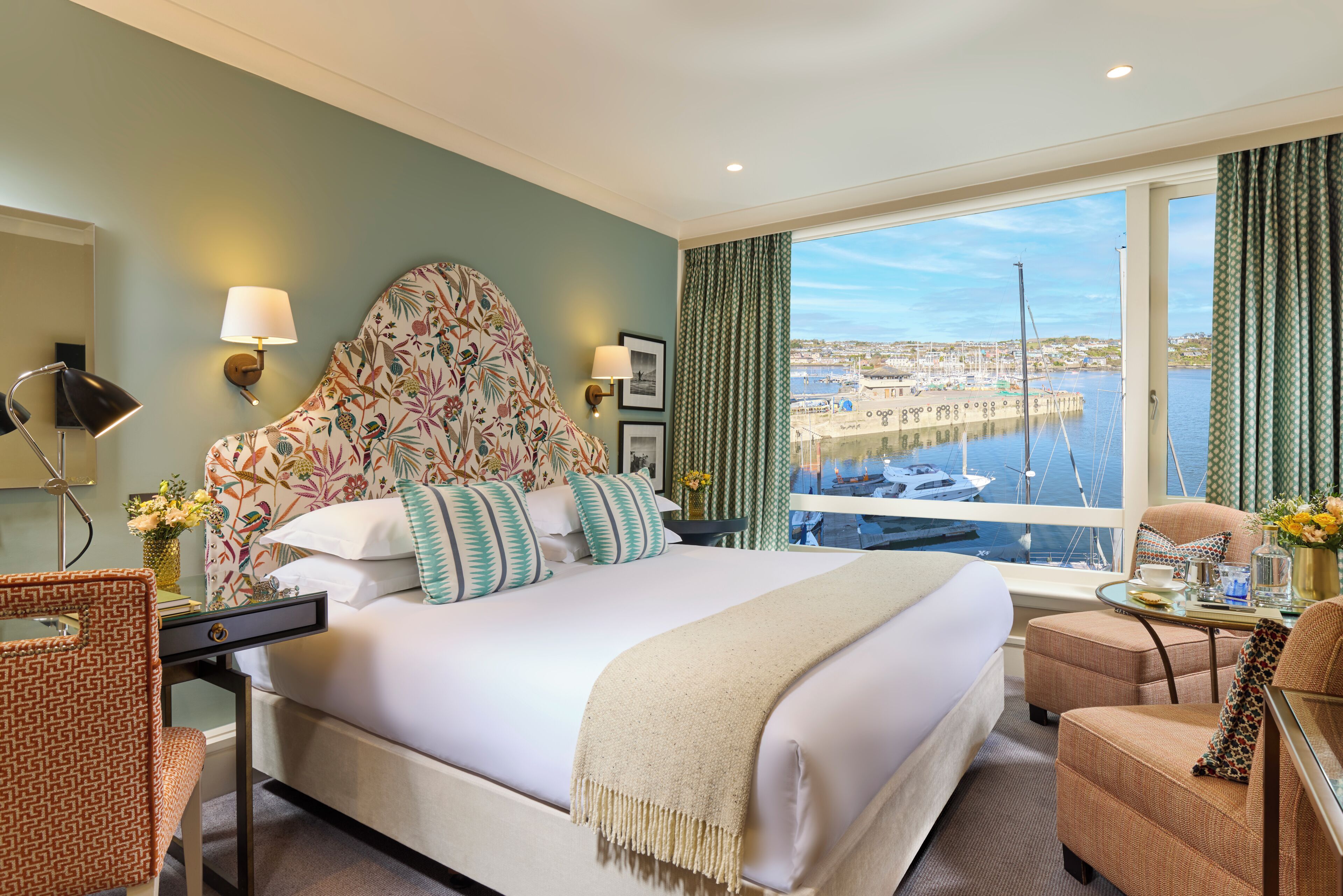 A hotel room with a large bed, a desk, a chair, a view of the marina, and a balcony with curtains.