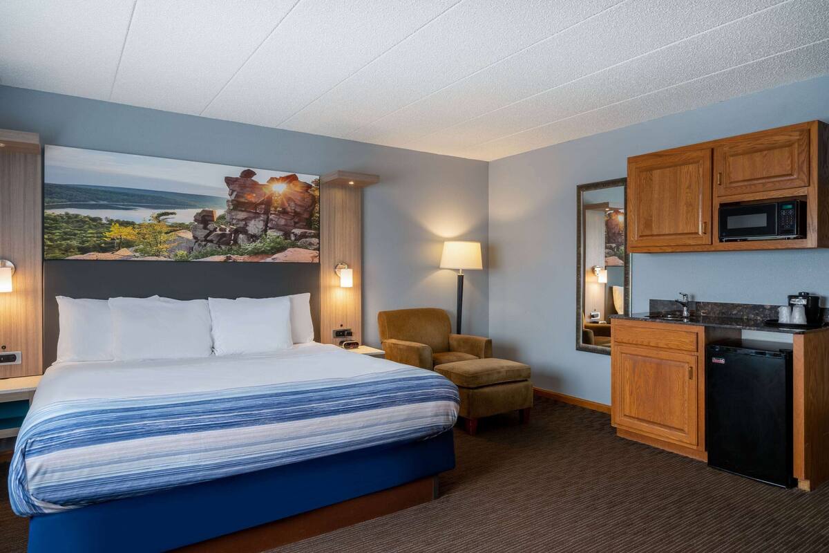 Habitación de hotel con una cama grande, un escritorio con microondas, una silla y un cuadro de un paisaje en la pared.
