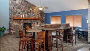 Een stenen openhaard, houten stoelen, een bar en een plafondventilator in een kamer met blauwe wanden en houten balken.