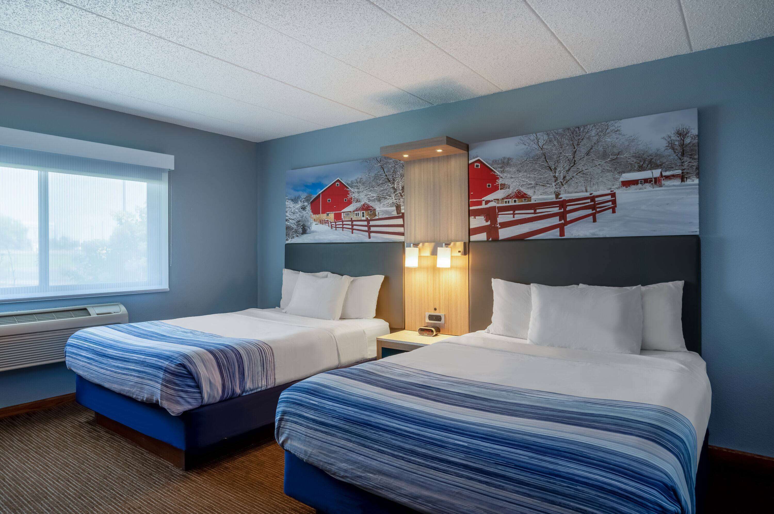 A hotel room with two beds, a large window, and a painting of a snowy farm scene.