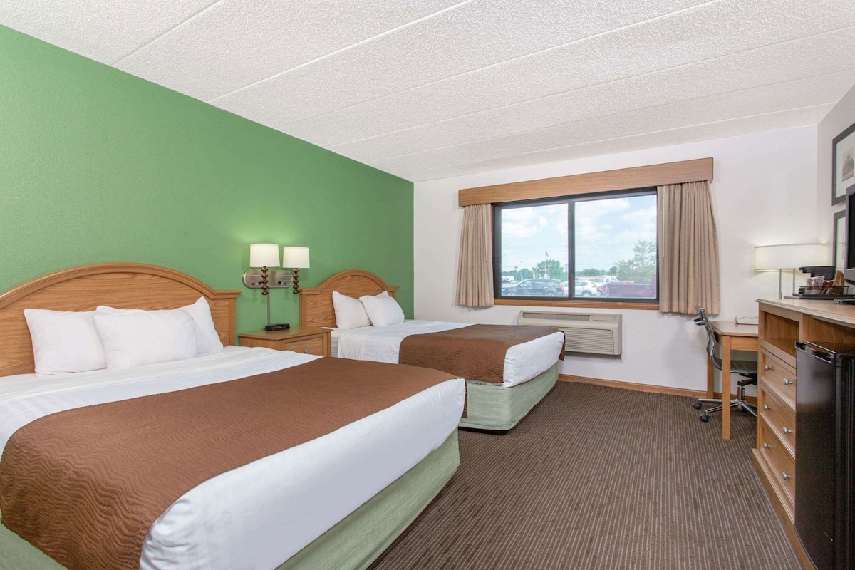 A hotel room with two beds, a desk, a chair, a window with curtains, and a view of the outside.