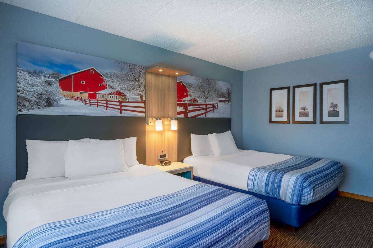Una habitación de hotel con dos camas, un cuadro grande de una escena invernal en el campo y tres fotografías enmarcadas en la pared.
