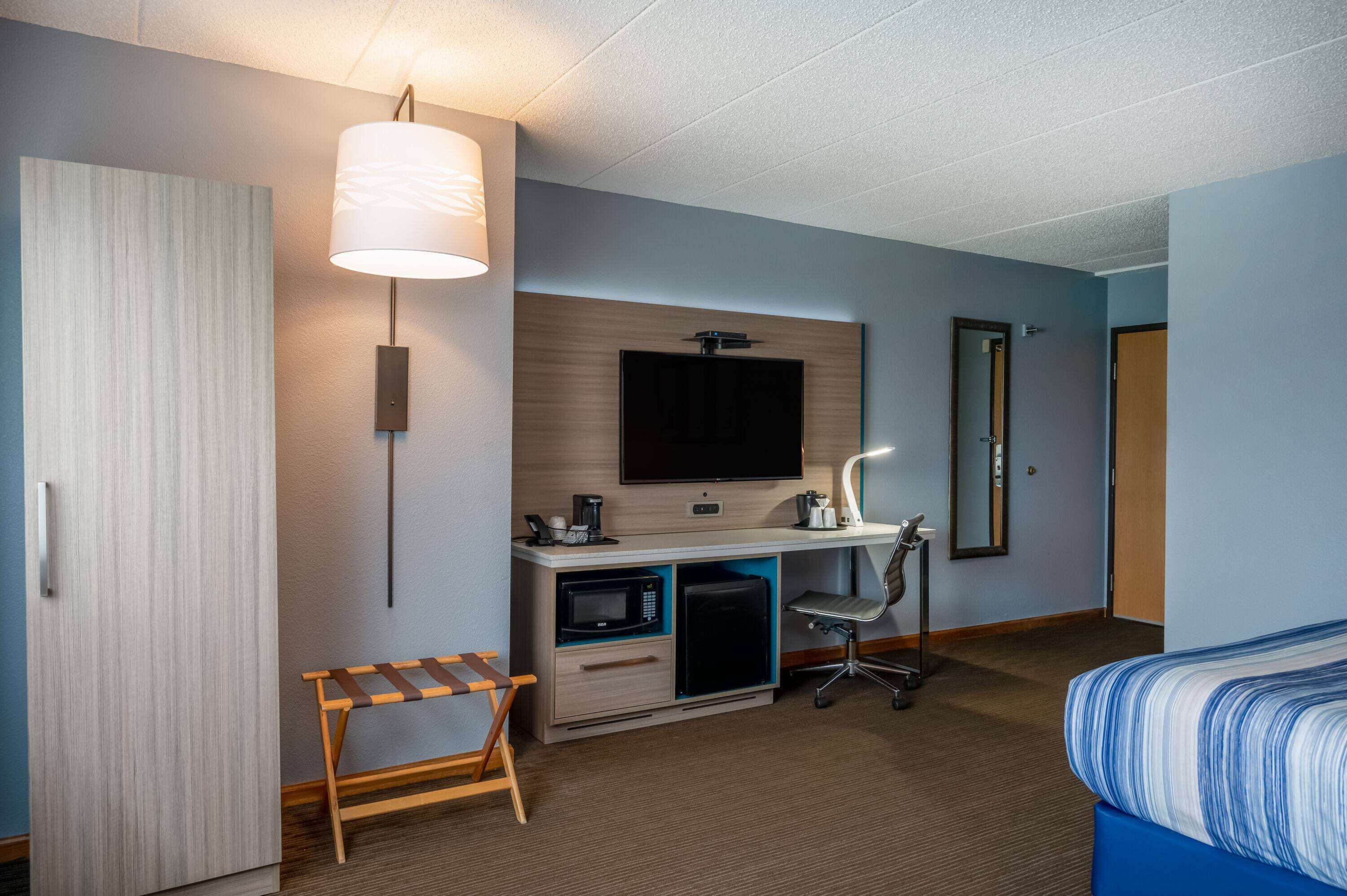 A hotel room with a bed, a desk with a computer, a microwave, a chair, and a TV.