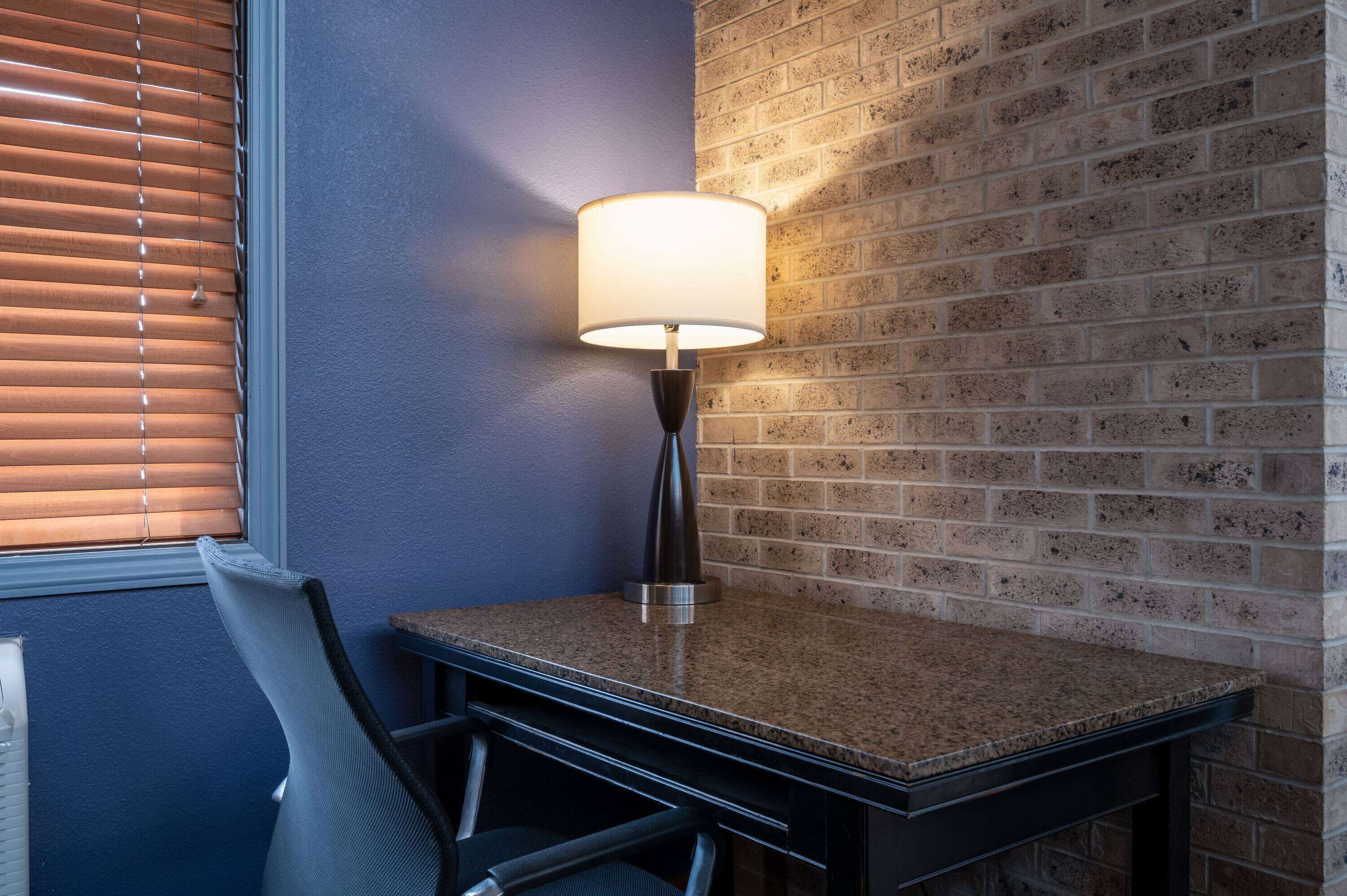 Una esquina de habitación con una mesa, una silla, una lámpara y un muro de ladrillo.