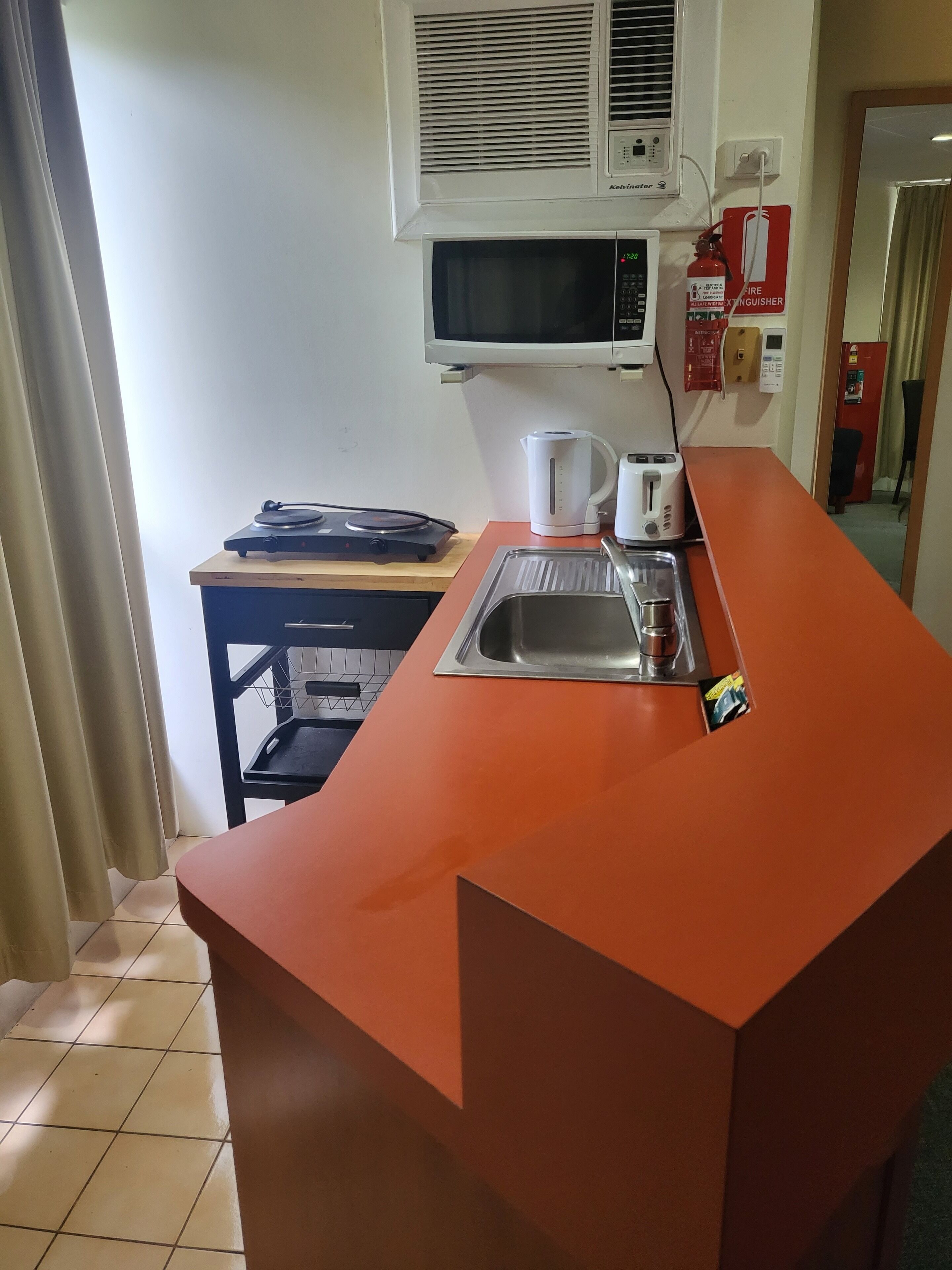 A kitchen with a microwave, kettle, toaster, and sink. There is a fire extinguisher and a small table with a kettle and a toaster.