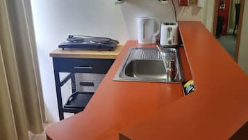A kitchen with a microwave, kettle, toaster, and sink. There is a fire extinguisher and a small table with a kettle and a toaster.