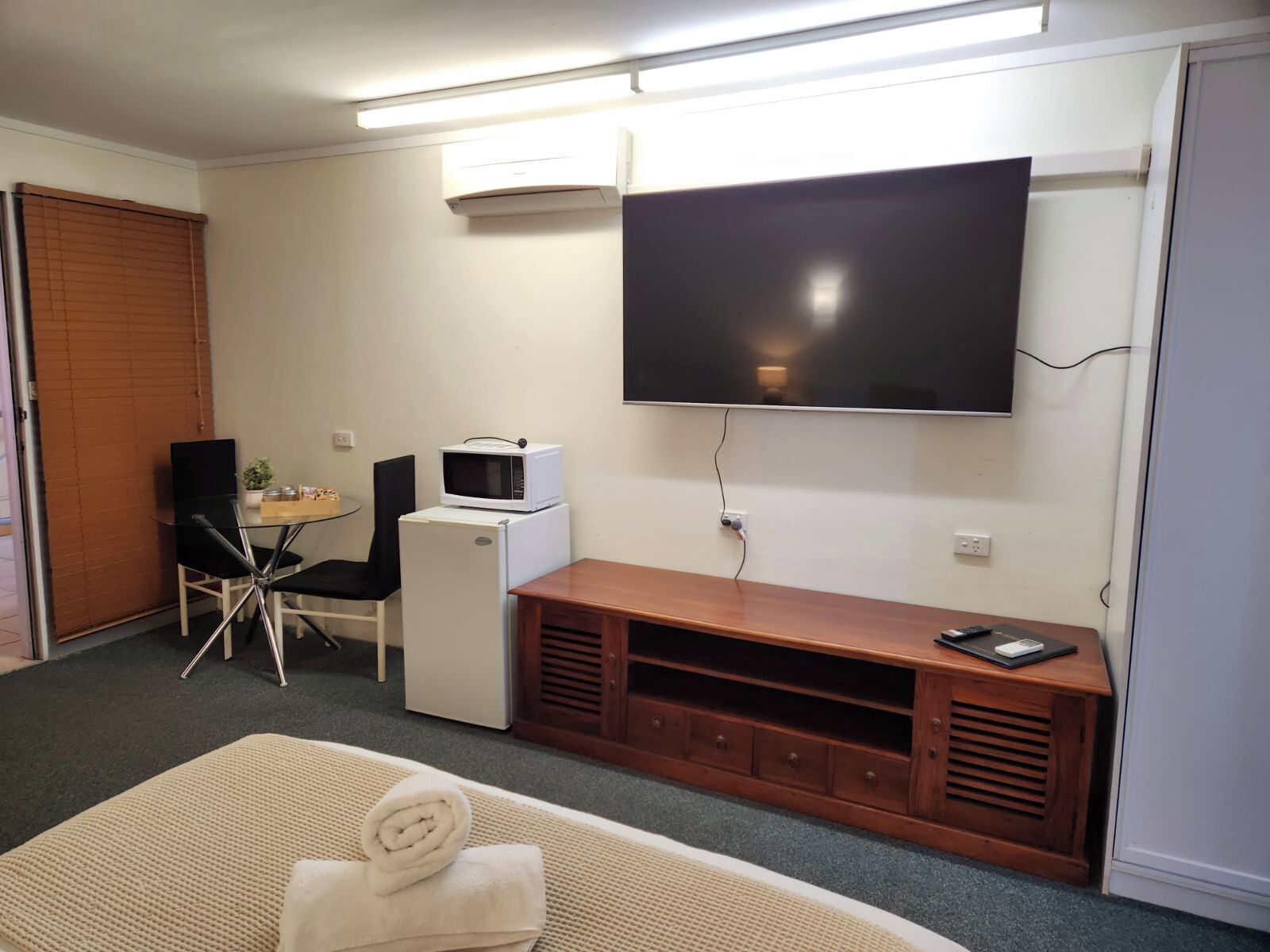 A compact room with a flat-screen TV mounted on the wall, a small kitchenette with a microwave, a wooden entertainment unit, and a small table with chairs.