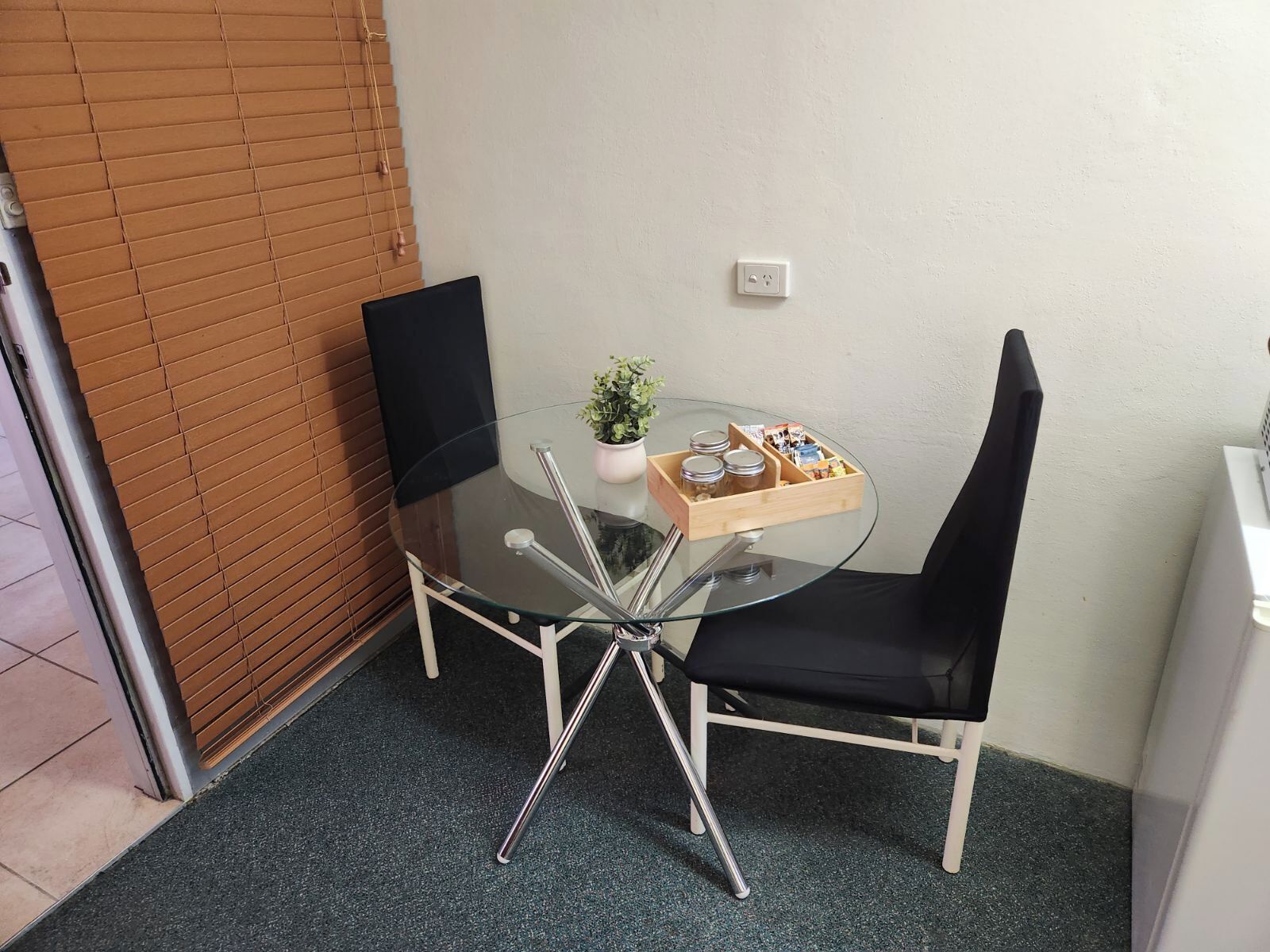 Una pequeña mesa de vidrio con una silla negra, una planta en maceta y un caja de madera con frascos y aperitivos.