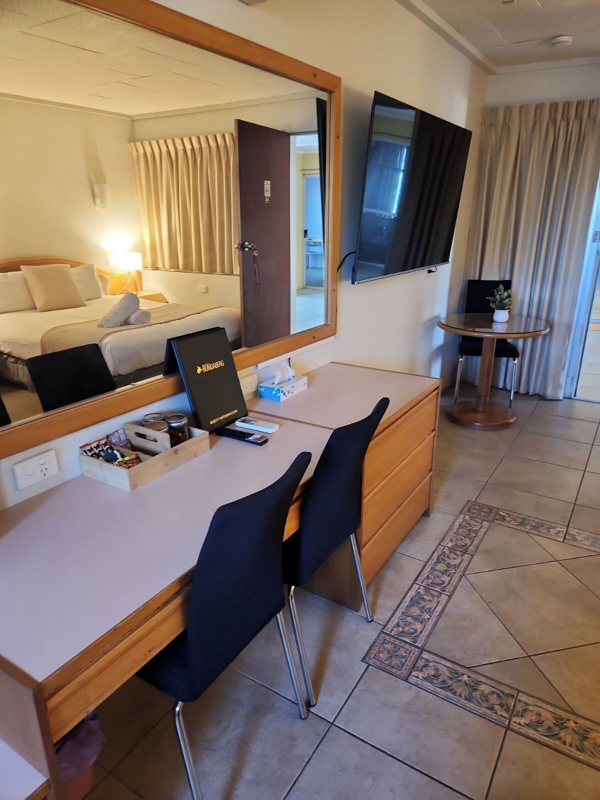 A hotel room with a bed, a desk with a computer, a chair, a TV, and a small table with a plant.