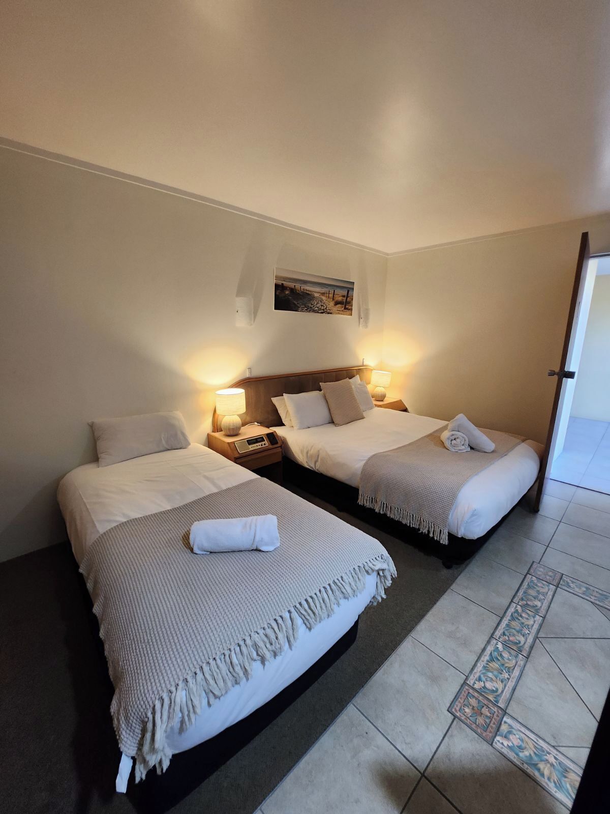 A hotel room with two beds, each with a bedside table and lamp, a small painting on the wall, and a tiled floor with a decorative border.