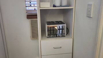 A compact kitchenette with a built-in toaster, a small trash bin, and a window with blinds.