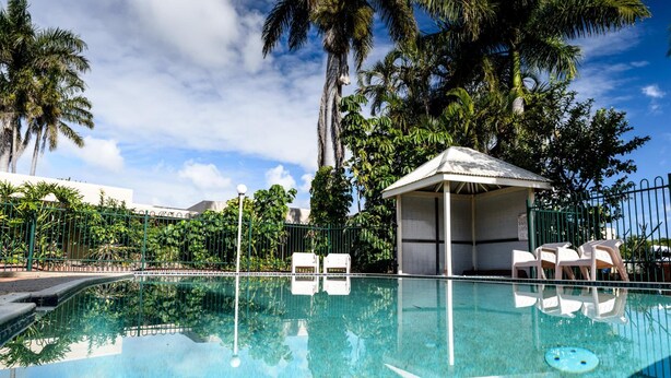 A clear swimming pool with lounge chairs and a gazebo, surrounded by palm trees and a fence. - Bundaberg International Motor Inn (Millbank)