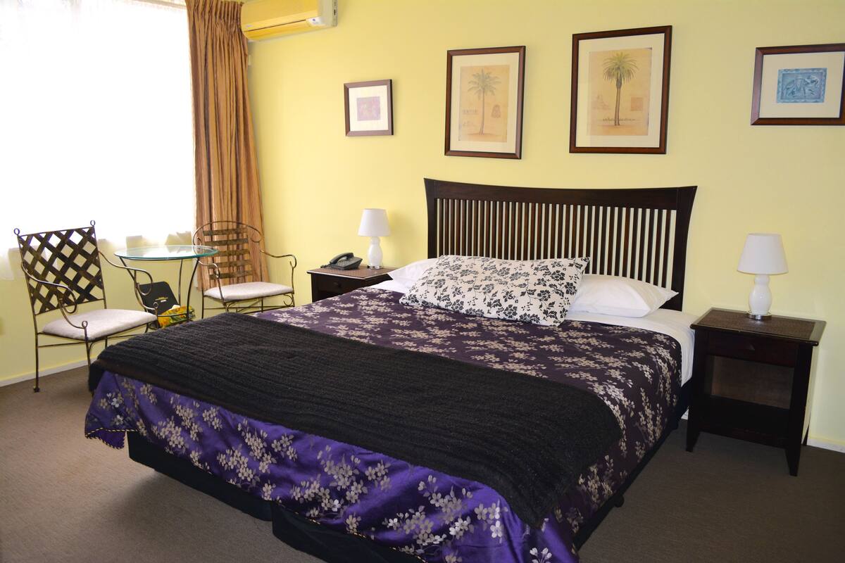 Habitación de hotel ordenada con una cama, dos sillas, una mesita y una mesita de noche con una lámpara.