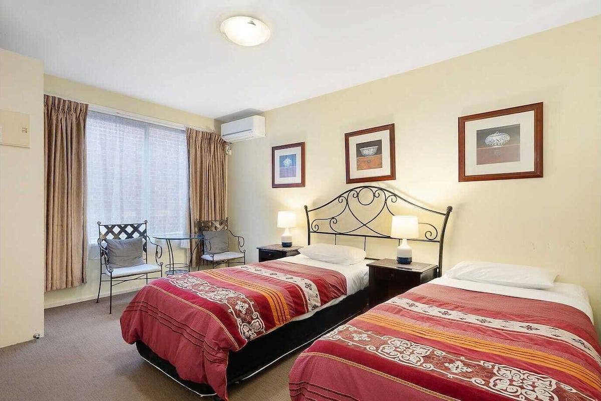 Habitación de hotel con dos camas, una mesita, una silla y un ventanal con cortinas.