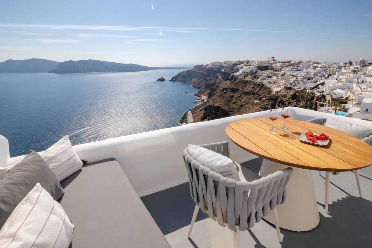 A terrace with a wooden table and white chairs, overlooking a coastal town with white buildings and a view of the sea.