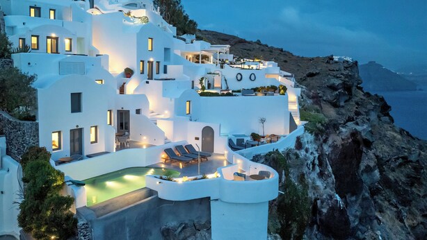 A white, multi-story building with a pool and lounge chairs, surrounded by rocky terrain and overlooking a body of water. - IKIES Santorini (Santorini)