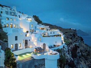 A white, multi-story building with a pool and lounge chairs, surrounded by rocky terrain and overlooking a body of water. - IKIES Santorini (Santorini)