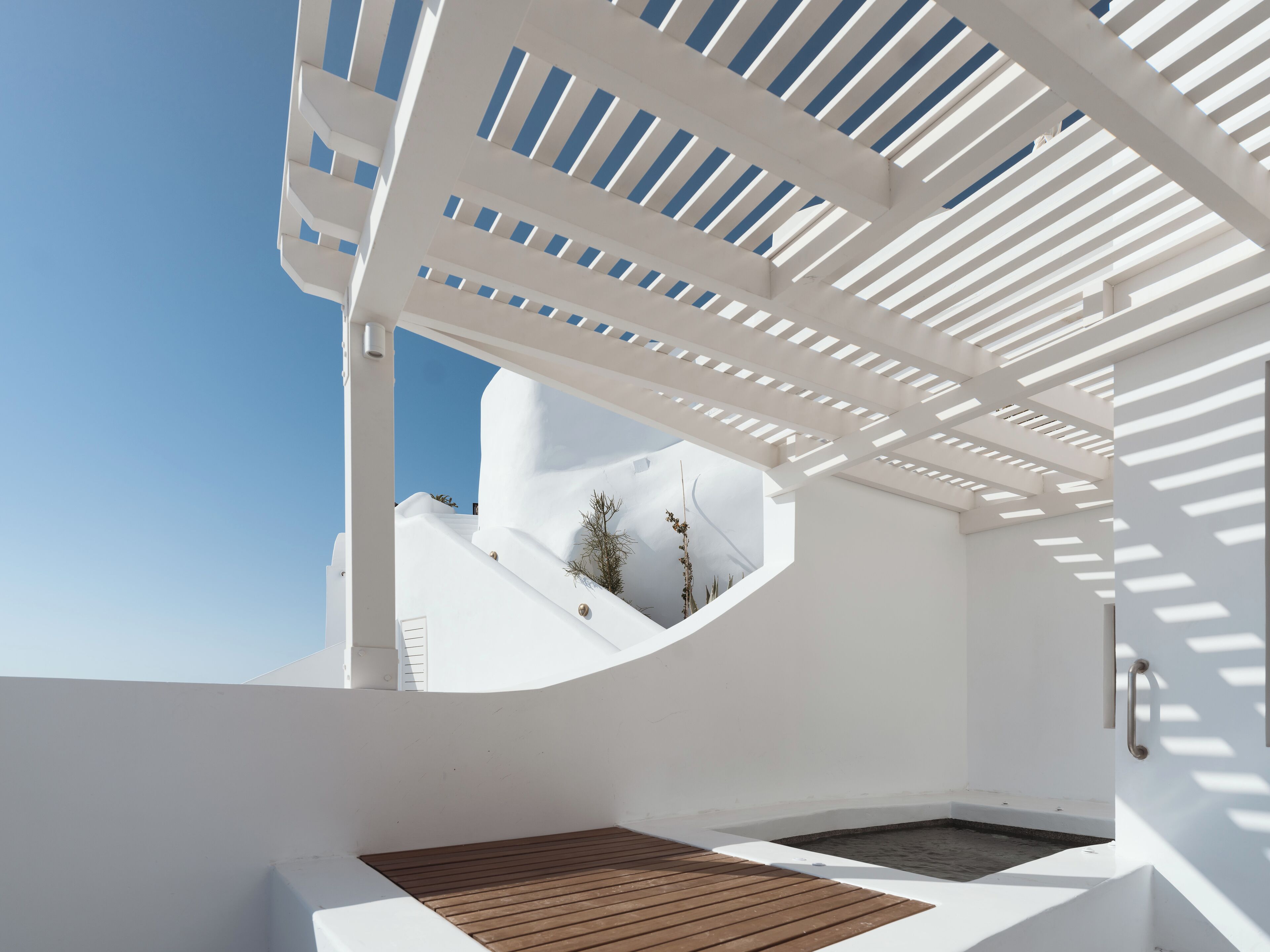 A modern outdoor deck with a white structure and a wooden floor, overlooking a white building and clear sky.