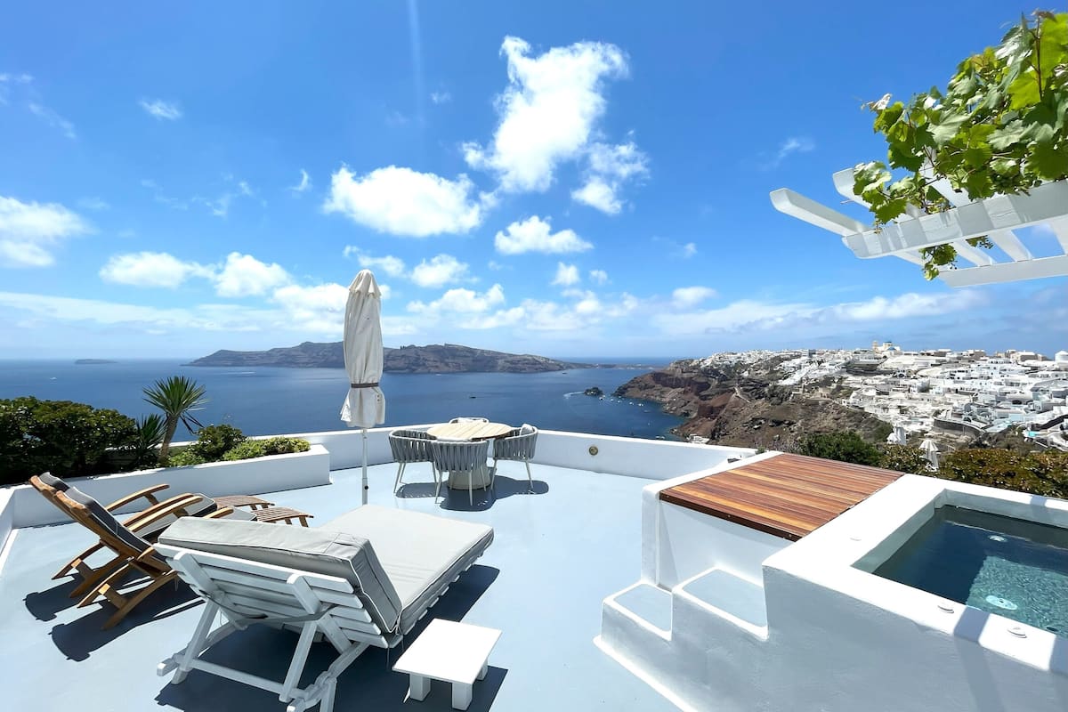 A sunlit outdoor seating area with lounge chairs and a table, overlooking a scenic coastal view with a pool and white architecture in the background.